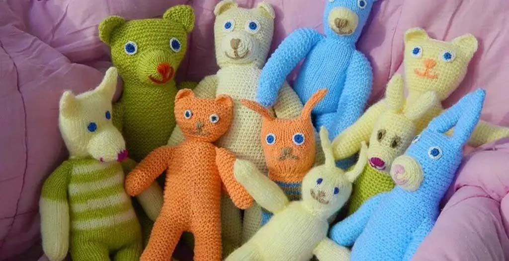 Group of colorful handmade knitted stuffed animals including bears, rabbits, and cats arranged on a pink blanket.