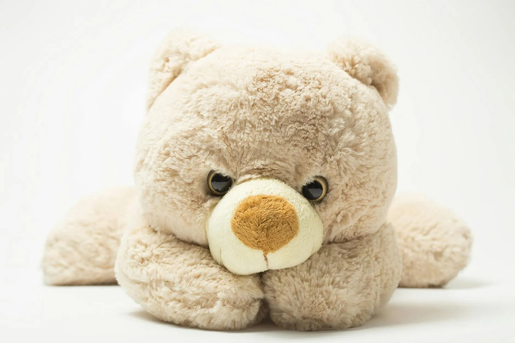 A fluffy beige teddy bear plush toy lying flat on a white background with its head resting on its paws, giving a cute and relaxed expression.