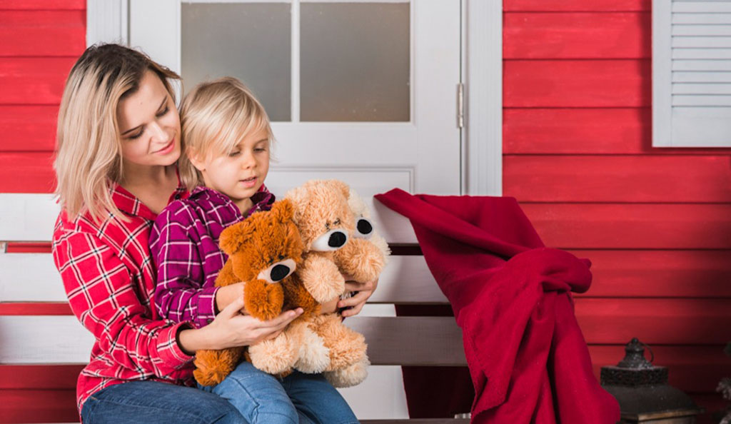 A mother and child sitting on a white bench outside a red wooden house, both dressed in plaid shirts, holding two soft teddy bears together.