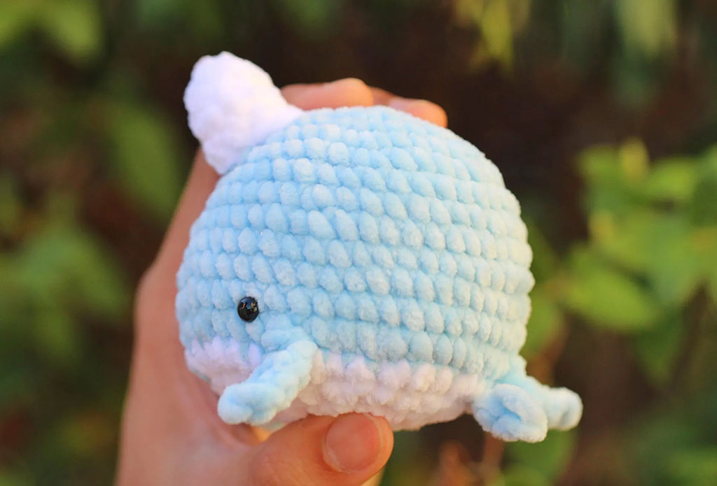 A hand holding a small, handmade blue whale plush toy with white details and soft bubble-textured fabric, set against a blurred green background.
