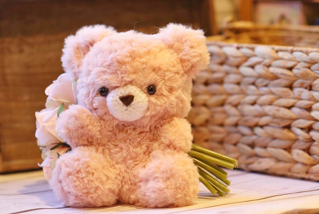 A soft pink teddy bear sitting on a wooden table holding a small bouquet of light pink flowers, with a wicker basket in the background.