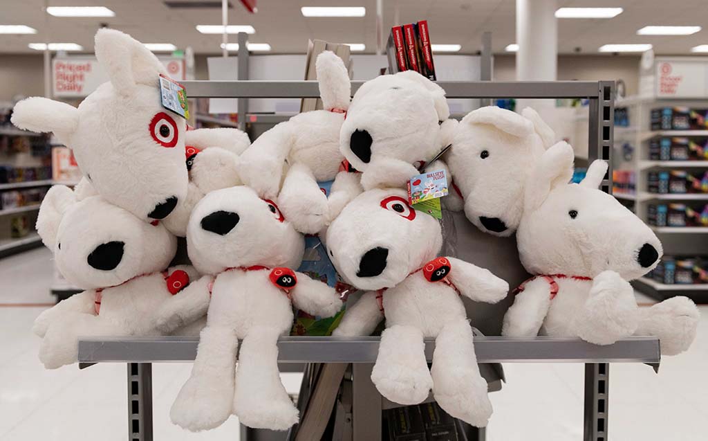 A store shelf filled with multiple white dog plush toys featuring red collars and red eye patches, neatly arranged in a playful pile inside a retail aisle.