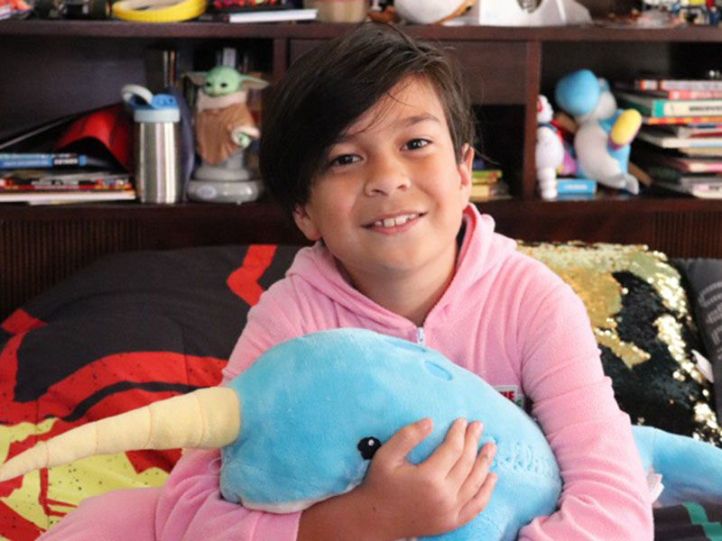 A smiling child in a pink hoodie sitting on a bed and hugging a large blue narwhal plush toy with a yellow horn.