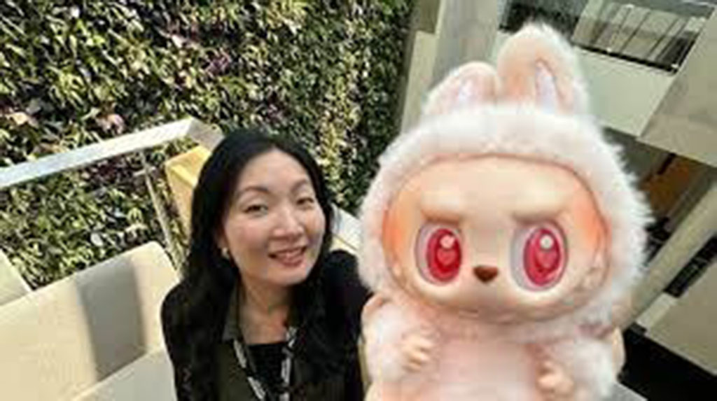 Smiling woman posing indoors while holding a large plush toy with big pink eyes and bunny ears, standing near a green wall background.