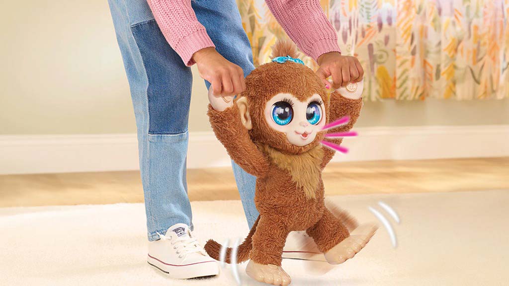 Child holding hands of an interactive monkey plush toy with big blue eyes, making it walk on the floor indoors.