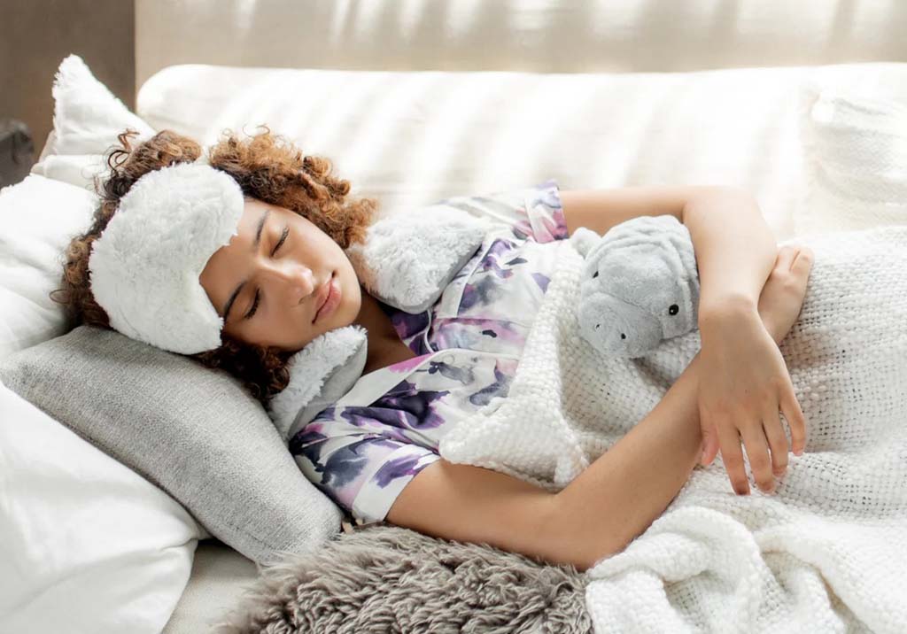 A woman wearing a soft sleep mask and floral pajamas rests peacefully on a bed while cuddling a small gray plush toy under a cozy white blanket.
