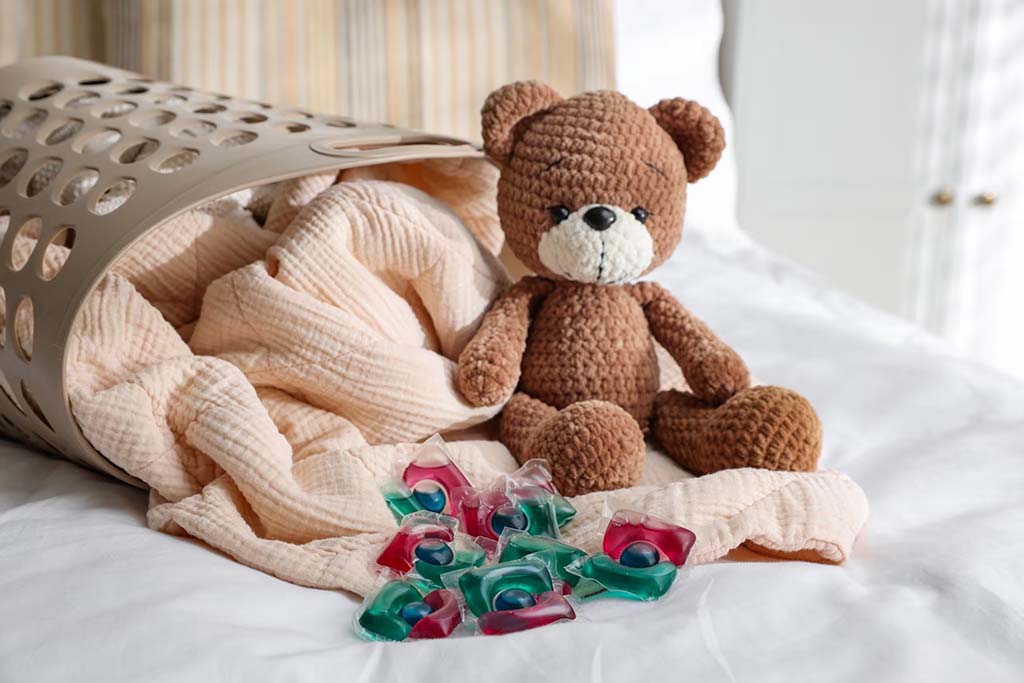 A knitted brown teddy bear sitting beside a laundry basket and blanket on a bed, with colorful detergent pods placed in front.