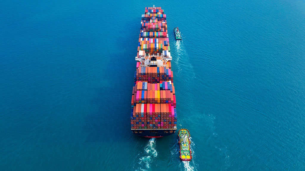Aerial view of a large cargo ship carrying colorful shipping containers across the ocean, accompanied by two smaller boats assisting in navigation.