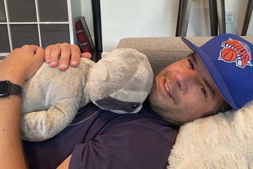 A man wearing a blue cap and navy shirt lying on a couch while hugging a soft gray plush toy, smiling comfortably.