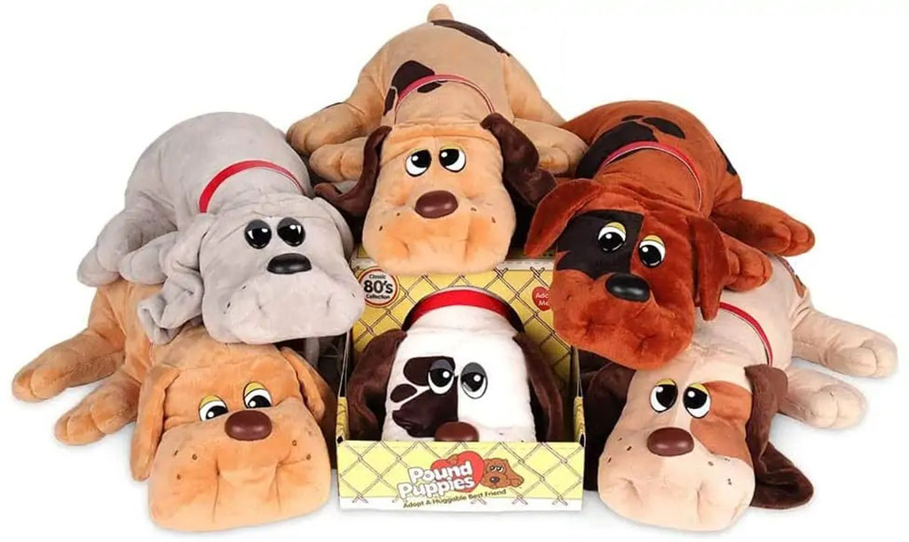 A group of vintage-style Pound Puppies plush toys in brown, tan, and gray colors stacked together, with one displayed in its original 80s-style yellow box.