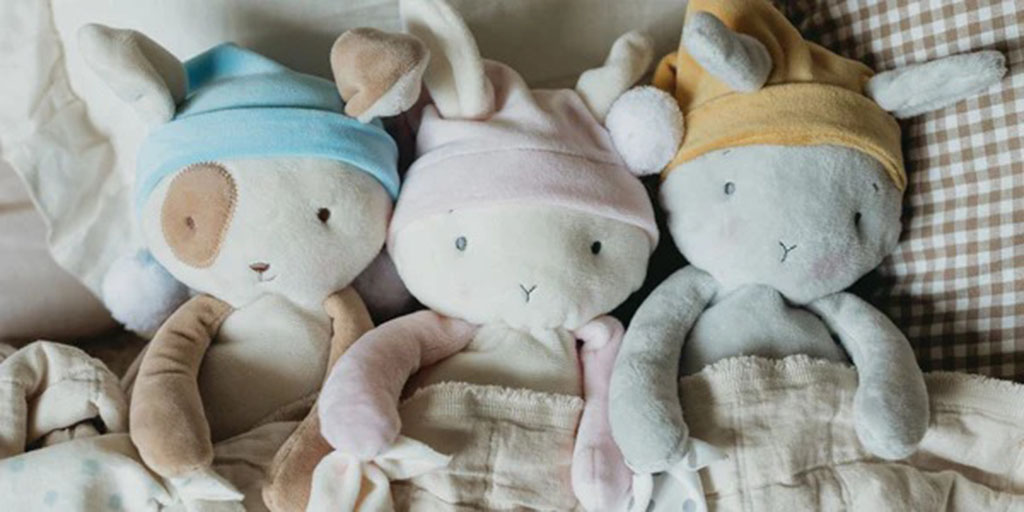 Three soft bunny plush toys wearing pastel nightcaps tucked under a cozy blanket on a bed.