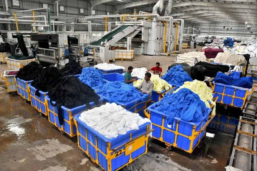 Workers sorting large bins of dyed fabric in a textile processing factory with automated machines and fabric rolls in the background.