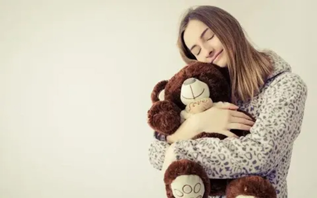 A woman wearing cozy pajamas hugging a soft brown teddy bear with a peaceful smile on her face.