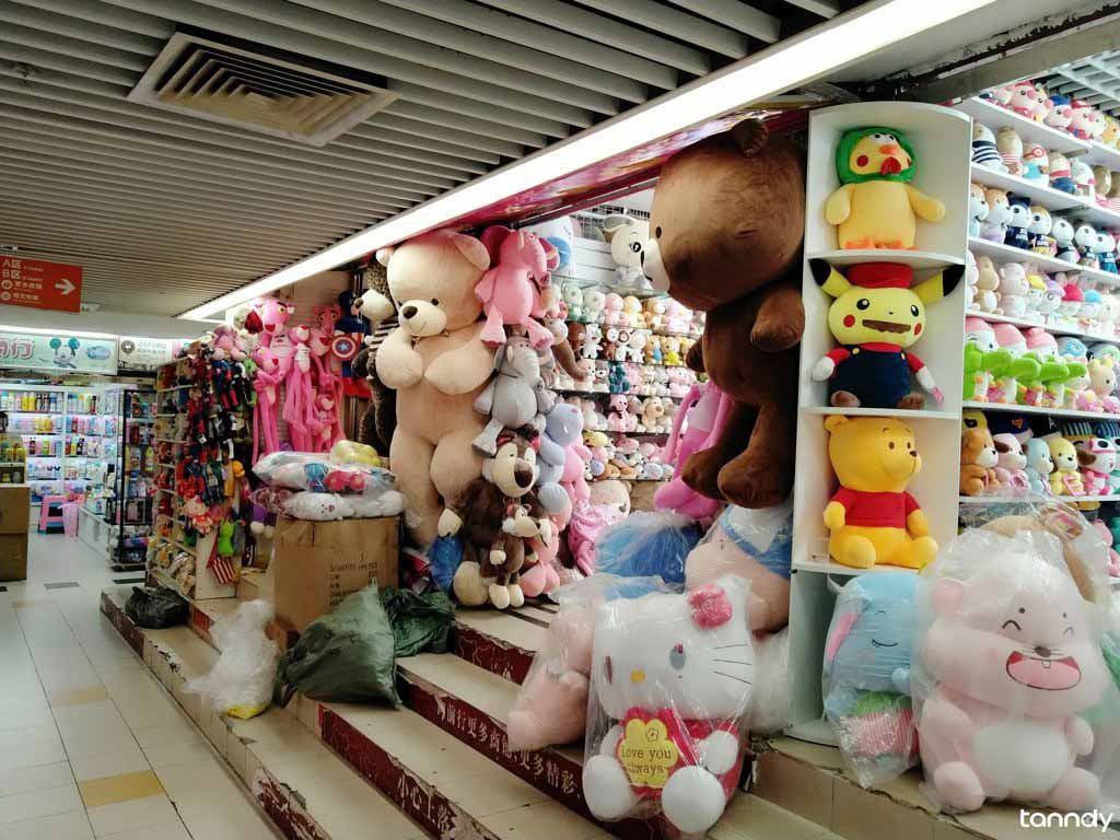 A wholesale toy shop filled with large and small plush toys, including teddy bears and cartoon characters, displayed on shelves and steps inside a brightly lit market.