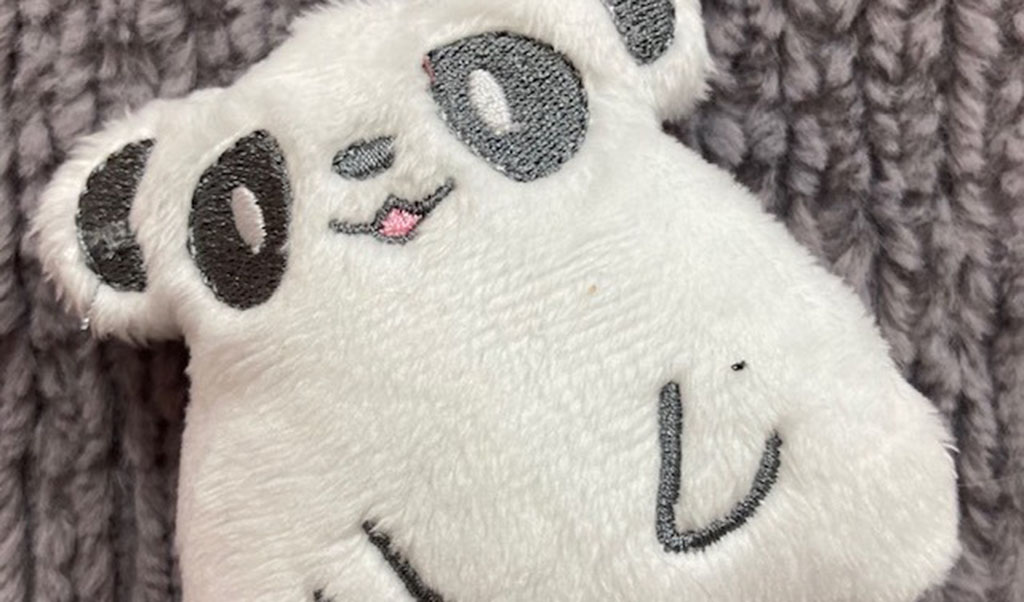 Soft white panda plush toy with embroidered black eyes and pink mouth on a gray knitted background.