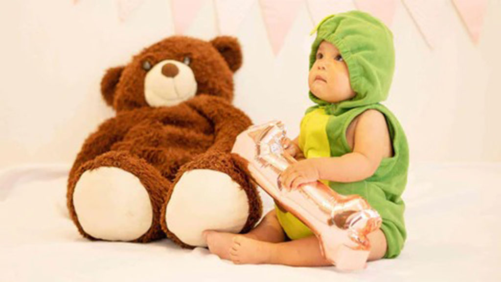 Baby dressed in a green costume holding a shiny balloon while sitting beside a large brown teddy bear on a white surface.