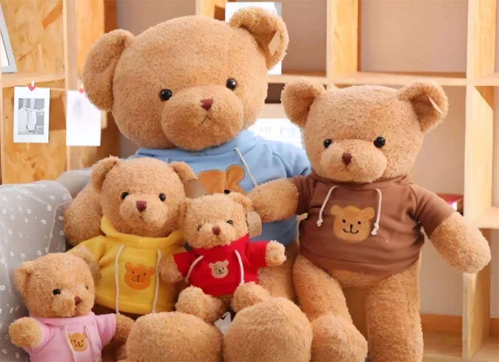 Group of cute teddy bears of different sizes wearing colorful hoodies, sitting together on a cozy couch in a warm indoor setting.
