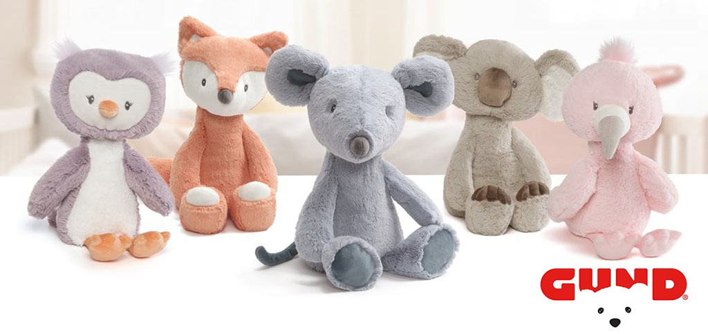 A lineup of soft GUND plush toys including a penguin, fox, mouse, koala, and flamingo displayed on a white surface with the GUND logo.