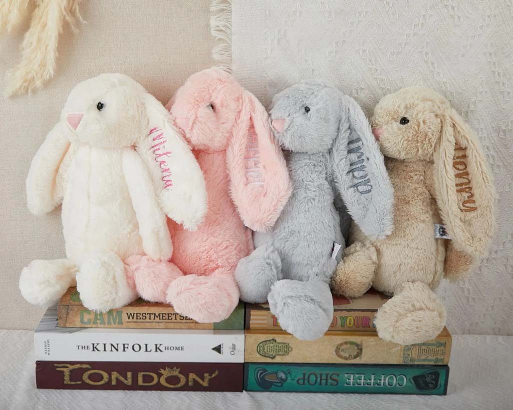 Four soft plush bunny toys in cream, pink, gray, and beige with embroidered names on their ears sitting on stacked books.