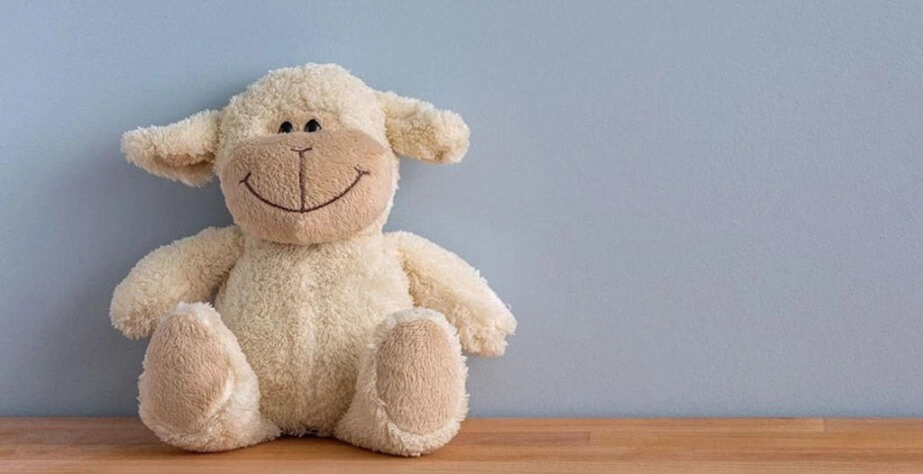 Soft cream-colored plush sheep with a big stitched smile sitting on a wooden surface against a light blue wall.