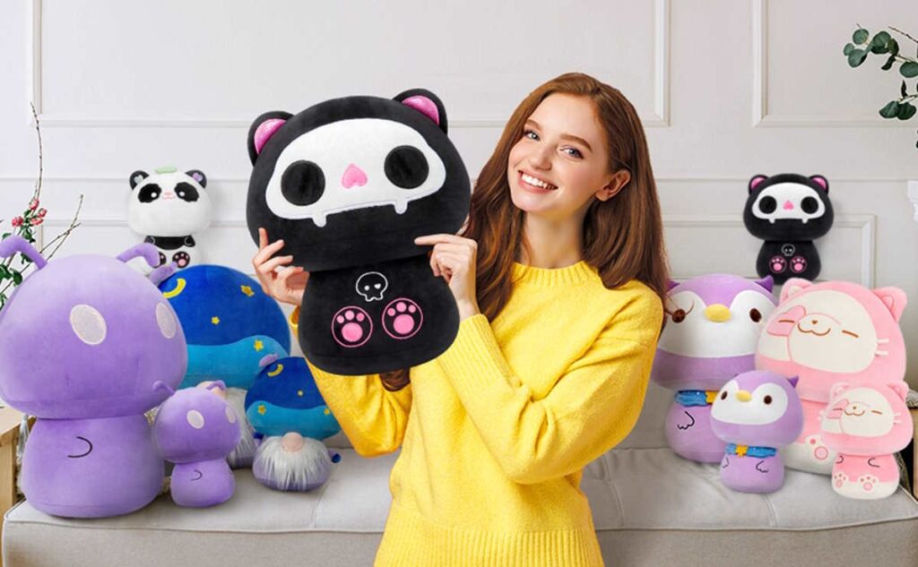 A smiling woman in a yellow sweater holds a black and pink skull-themed plush toy, surrounded by colorful animal and mushroom plushies on a sofa.