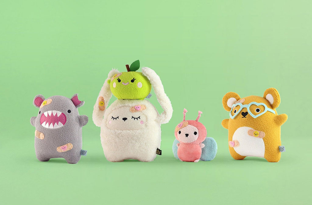 Colorful set of small plush toys in pastel colors including a monster, bunny, bug, and bear with bandage decorations on a light green background.