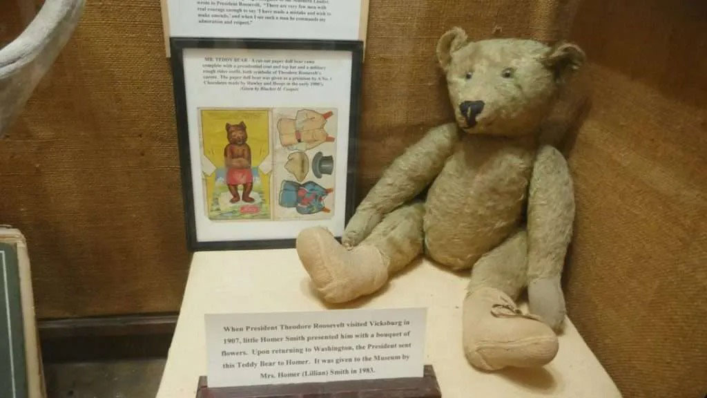 An old teddy bear displayed in a museum exhibit with historical notes and vintage illustrations explaining its connection to early teddy bear history.