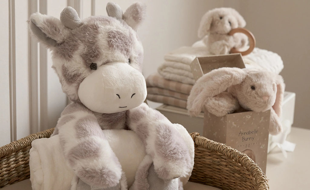 Soft pastel plush toys including a giraffe and bunnies arranged in a cozy nursery setting with blankets and gift boxes.