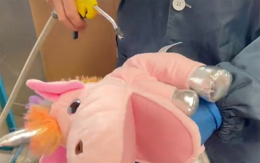 Factory worker using an air blower to remove dust from a pink unicorn plush toy during quality inspection.