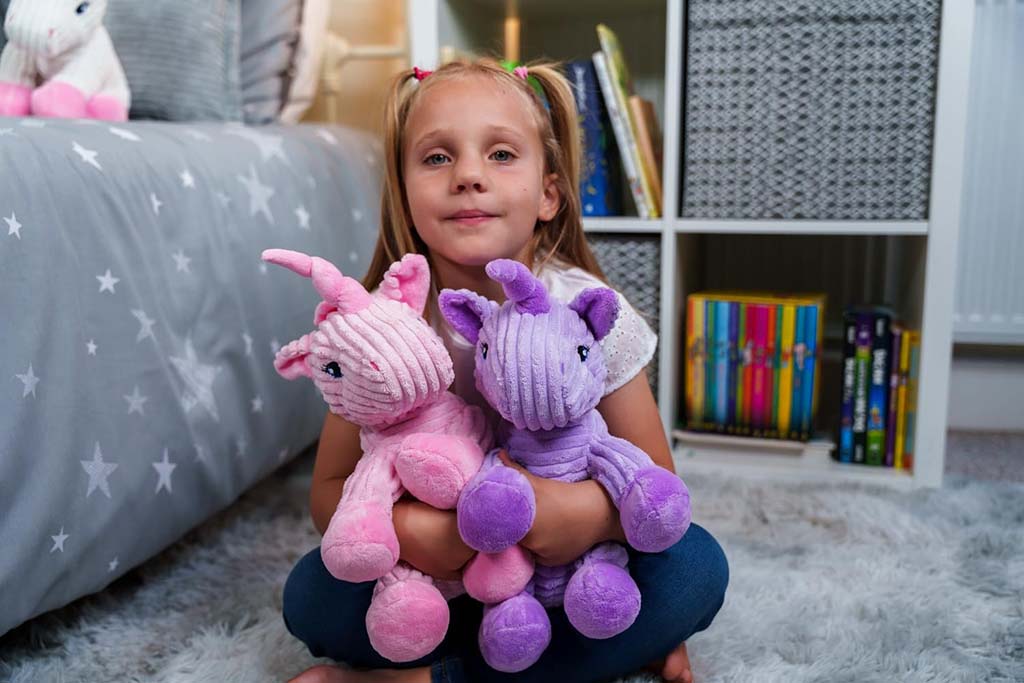 A young girl sits on a soft carpet in her bedroom, smiling and hugging two plush unicorn toys, one pink and one purple.