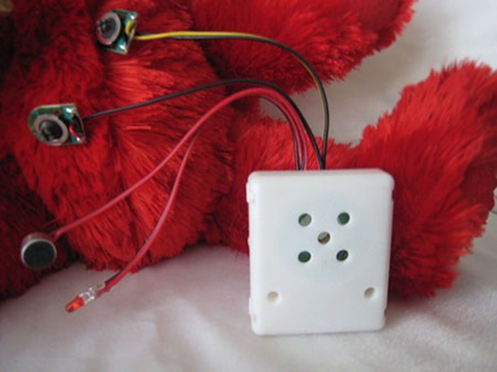 White electronic sound module with wires attached, shown next to a red stuffed animal for toy integration.