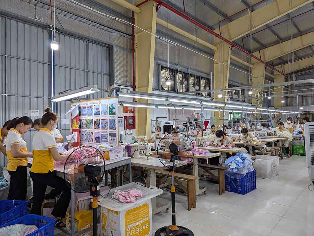 Workers in yellow uniforms sewing and assembling plush toy fabrics in a clean, organized workshop with bright lighting and industrial fans.