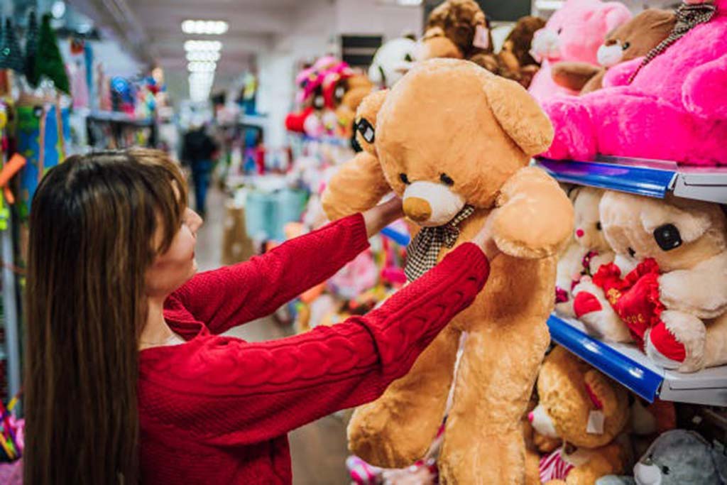 A woman in a red sweater holds a large brown teddy bear while browsing plush toys on store shelves filled with colorful stuffed animals.