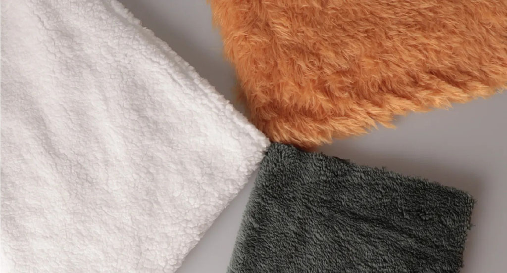 Three fabric swatches showing various plush materials — white sherpa fleece, orange long-pile faux fur, and dark gray short-pile minky — used in stuffed toy production.