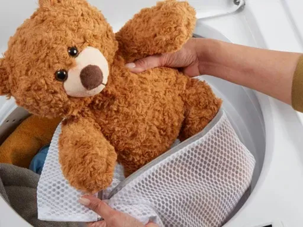 Person placing a brown teddy bear into a mesh laundry bag before washing it in a washing machine to protect its fur and stitching.