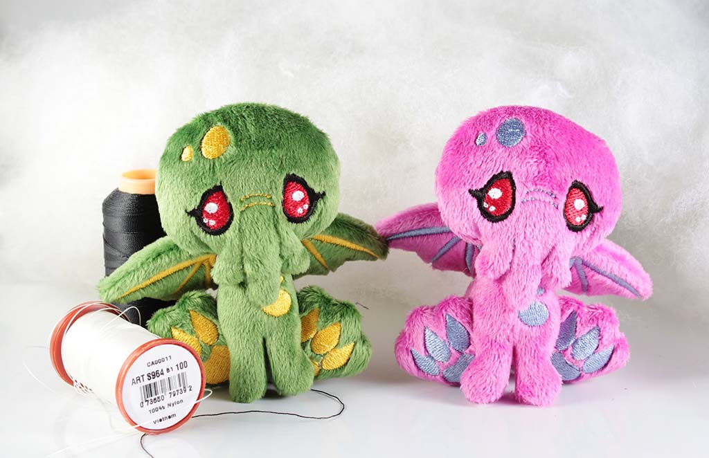Green and pink fantasy octopus plushies with embroidered red eyes and wings, placed beside sewing threads on a white background.