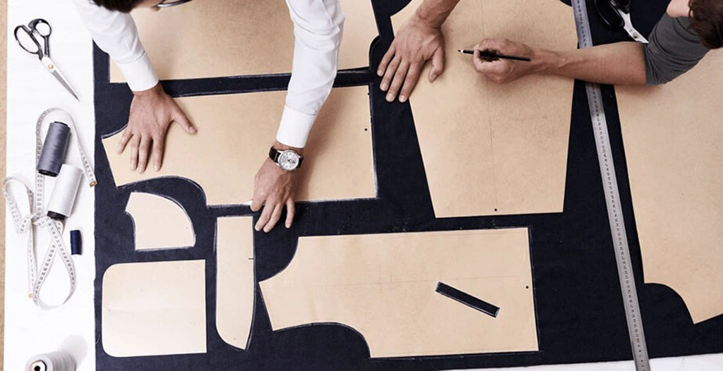 Designers working on cardboard templates for plush toy pattern making, surrounded by fabric, scissors, measuring tape, and sewing tools on a worktable.