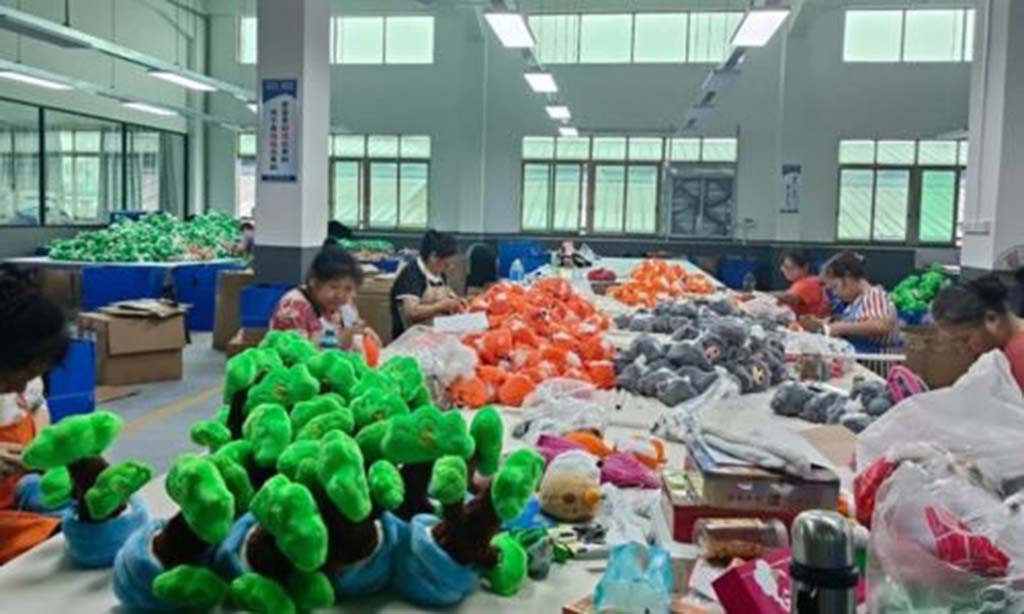 Workers in a toy factory assembling colorful plush toys, including green, orange, and gray stuffed figures, on long production tables.