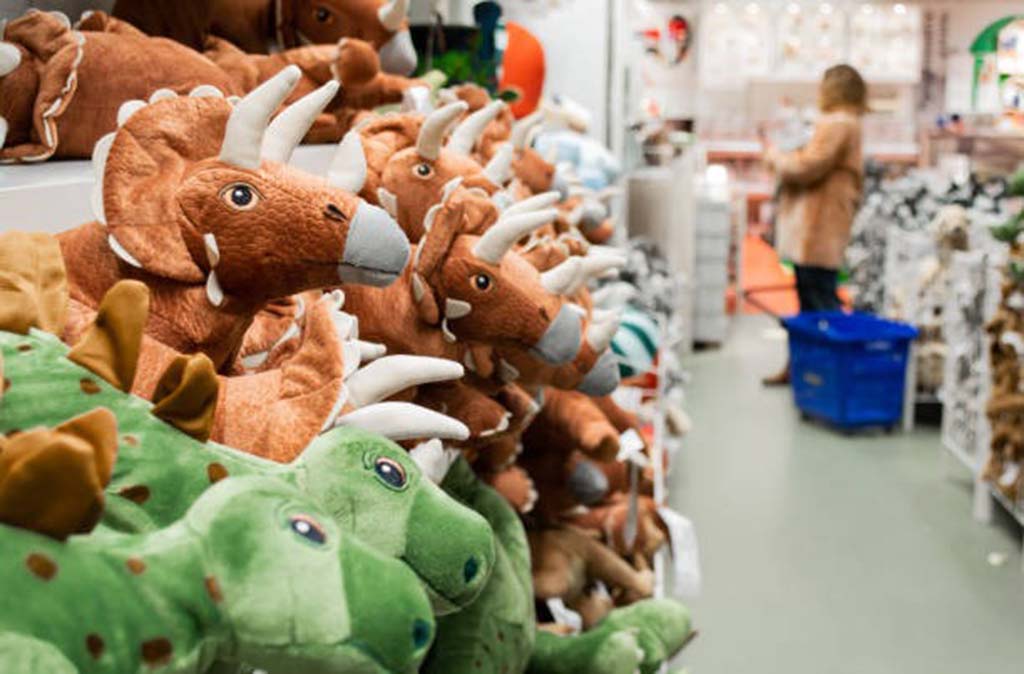 Shelves filled with soft dinosaur plush toys in green and brown colors displayed in a toy store, with a shopper browsing in the background.