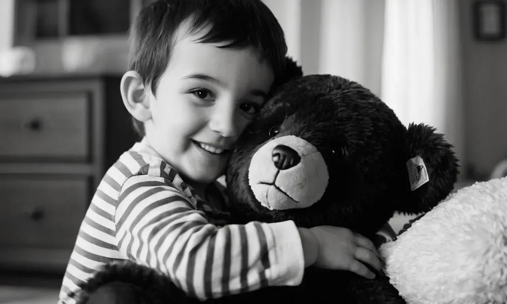 A smiling young boy in a striped shirt hugging a large teddy bear in a cozy home setting, captured in black and white.