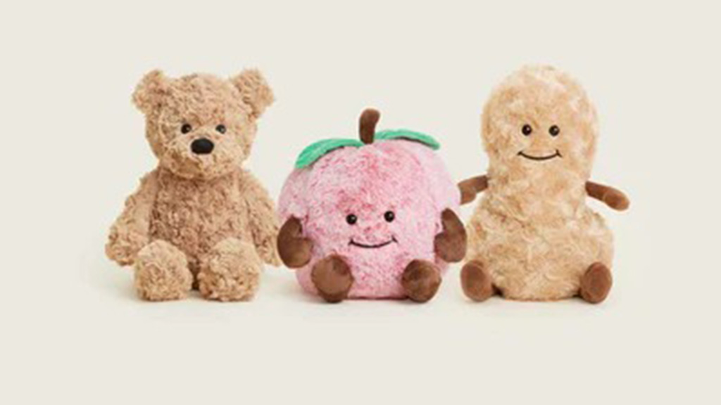 Three adorable plush toys — a teddy bear, a pink fruit-shaped plush with green leaves, and a peanut-shaped plush — all smiling and sitting side by side on a light beige background.