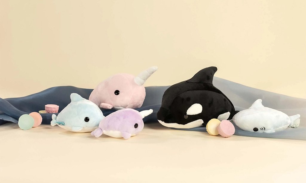 Two smiling plush cow toys, one blue and one pink, sitting on green grass with colorful cartoon tags attached to them.