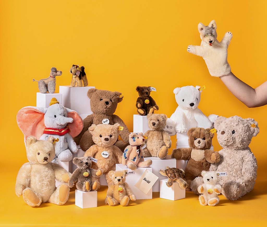 A display of Steiff plush animals and teddy bears, including elephants, dogs, and bears, arranged on white blocks against a yellow background.