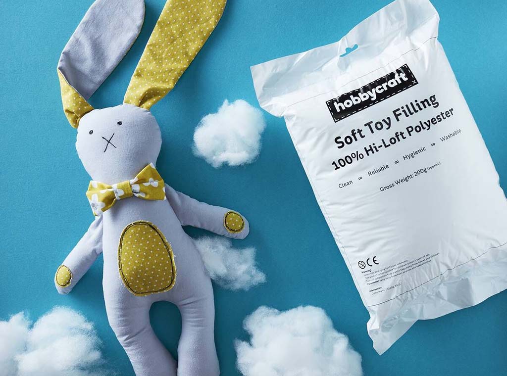A handmade bunny plush toy beside a bag of 100% Hi-Loft polyester stuffing on a blue background with fluffy fiber clouds.