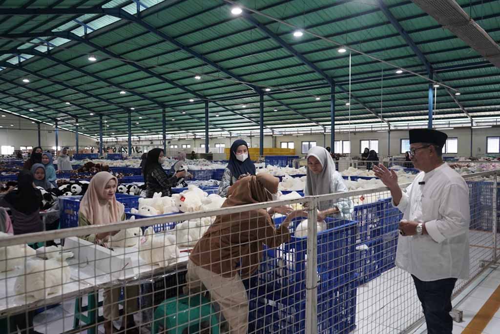 Workers in a large, well-lit factory assemble and inspect white plush toys, while a supervisor in a white shirt oversees the production process.