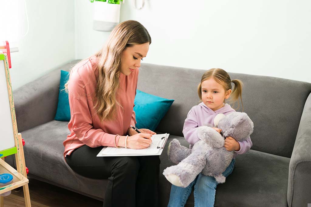 Young girl sitting on a couch holding a gray teddy bear while a therapist takes notes, illustrating the comforting role of plush toys in therapy sessions.