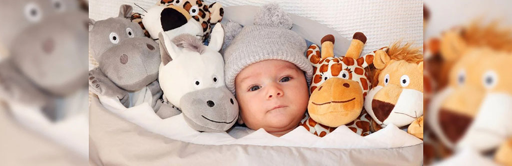 Baby wearing a gray hat lying in bed surrounded by soft plush animals including a giraffe, lion, and hippo.