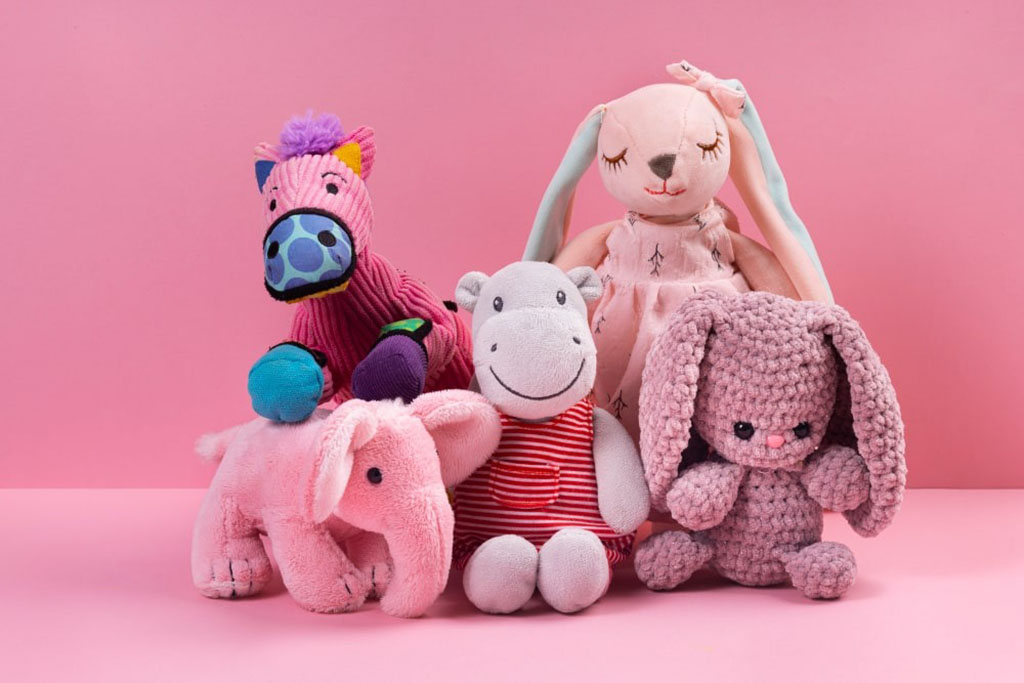 Group of adorable plush toys including a pink elephant, purple zebra, gray hippo, crocheted bunny, and a soft rabbit doll with long ears, all arranged on a pink background.