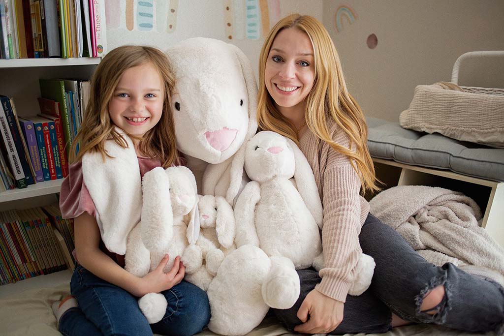 Smiling mother and daughter sitting on the floor together, surrounded by large and small white bunny plush toys in a cozy room with bookshelves and soft blankets.