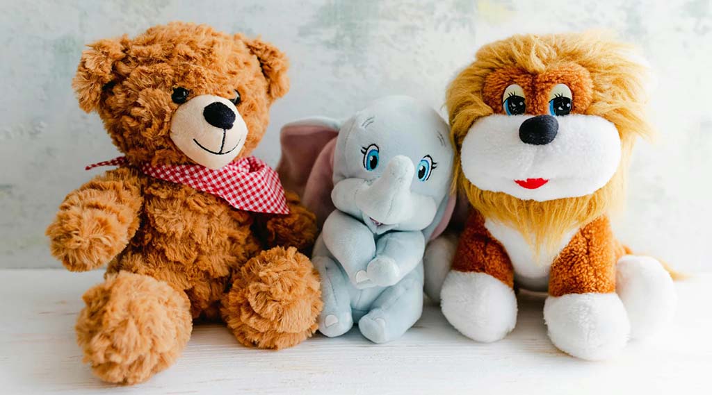 Plush teddy bear, elephant, and lion sitting together on a white surface showing different stuffed toy designs.